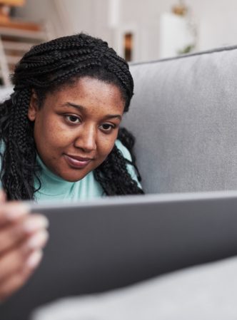 Portrait of curvy African American woman using laptop while lying on couch and relaxing at home in minimal interior, copy space