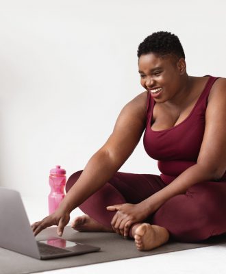 Positive overweight african american young woman in sports clothes sitting on fitness mat in front of laptop, enjoying remote workout while staying home during coronavirus pandemic, copy space